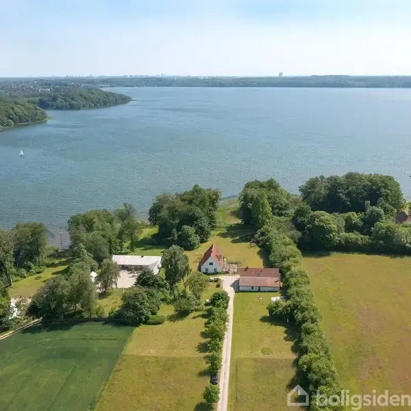 Hvid bygning ved en vej omgivet af grønne marker og træer, nær sø med klart vand og blå himmel. Baggrund indeholder mere skov og vand. Ingen synlig tekst.