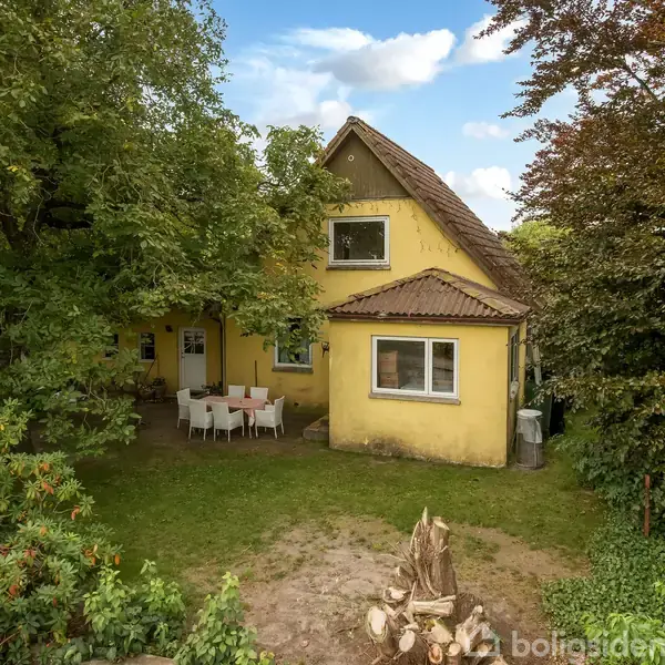 Et gulmalet hus med en spids gavl står omgivet af tætte, grønne træer. Foran huset ses et bord med stole placeret på en græsplæne.