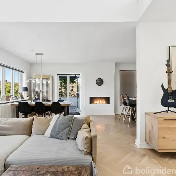 Sofa i forgrunden med puder, guitar på kommode til højre, åben stue med spisebord og stole. Store vinduer til venstre lader lys ind, pejs i baggrunden.