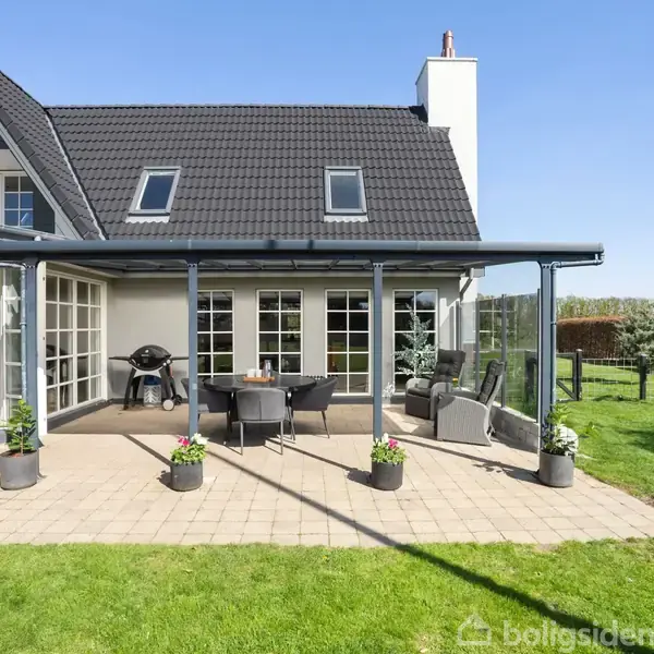 Hus med sort tag og store vinduer, åbne døre til en terrasse med møbler og grill. Omgivet af græsplæne og klar himmel.