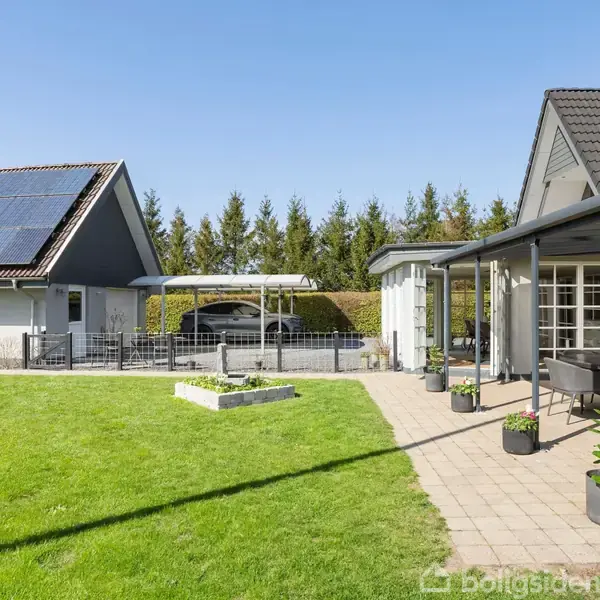 Et moderne hus med solpaneler på taget står i en have med græsplæne og flisebelagt terrasse. I baggrunden er der en carport og en hæk.
