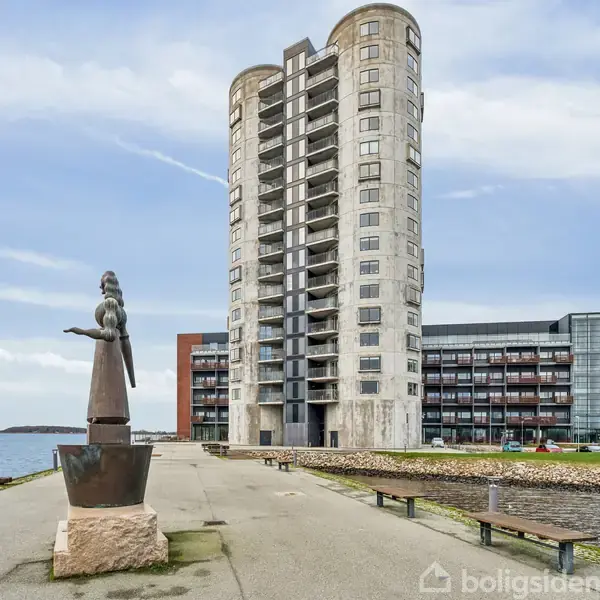 En bronzestatue af en siddende figur kigger ud over vandet på en havnepromenade. En høj, cylinderformet bygning står i baggrunden med moderne lejlighedskomplekser langs kajen. Skilt viser "PFA Kollegiet".