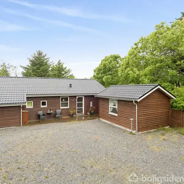 En træbeklædt husbygning med gråt tag står omgivet af grønne træer. Foran huset er der en grusbelagt forplads med nogle potteplanter ved indgangen.