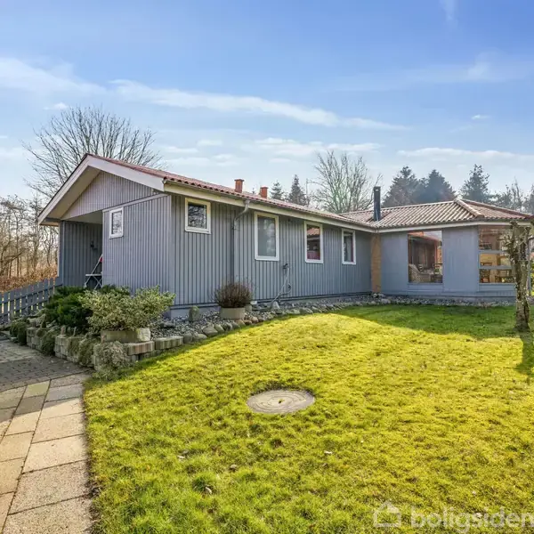 Gråt træhus står stille i en solrig have med græsplæne. Flisebelagt sti leder til indgangen, omgivet af buske og træer i baggrunden.