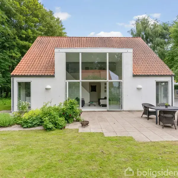 Et hvidt hus med rødt tegltag har store glasdøre, der åbner ud mod en terrasse med havemøbler. Huset ligger i en grøn have omgivet af træer.