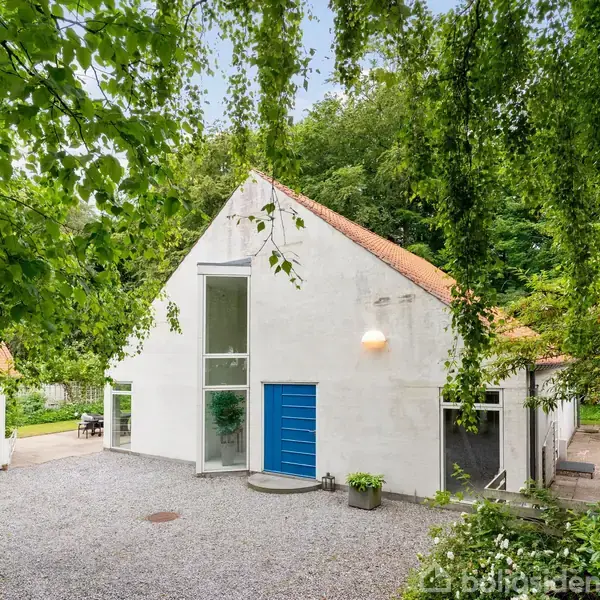 Et hvidt, moderne hus med blå dør står på en grusbelagt gårdsplads. Det er omgivet af grønne træer og buske, med en garage til venstre.