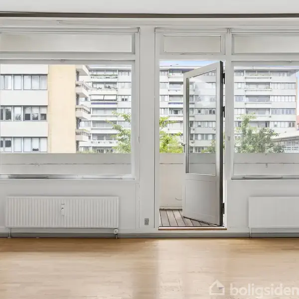 En åben altandør fører ud til en balkon med en udsigt over nabobygninger. Rummet har trægulv og radiatorer under vinduerne. Store vinduer slipper naturligt lys ind.