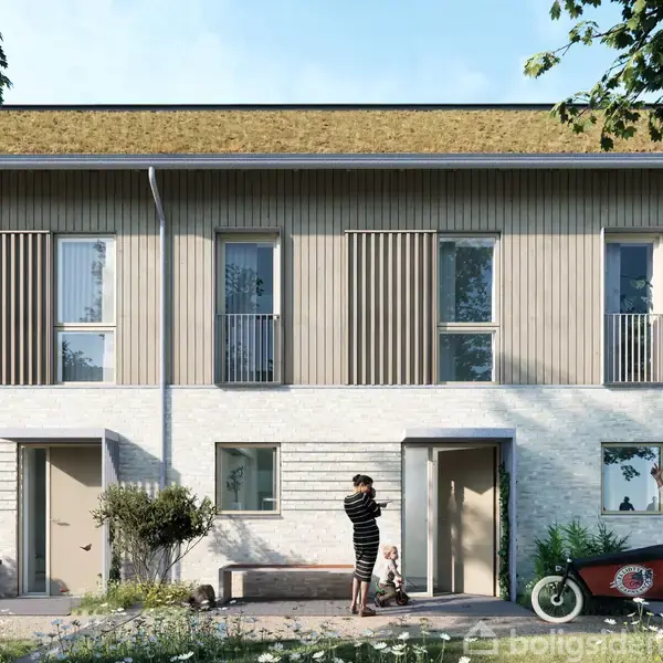 To-etagers rækkehus med moderne design og græstag. To personer står foran bygningen; én holder et barn, den anden skubber en cykel. Omgivet af græs og planter i sollys.