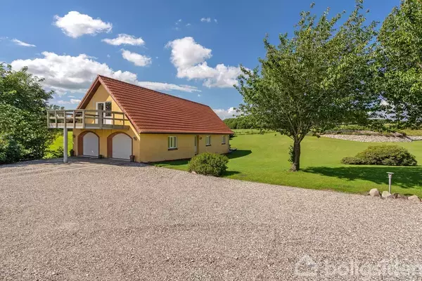Et gult hus med to garageporte står på en grusbelagt plads. Ved siden af er en grøn græsplæne med et stort træ under en klar blå himmel med skyer.