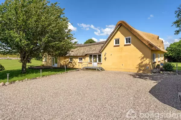 Et gult stråtækt hus med en stor grusbelagt gårdsplads foran. Til venstre står et træ, og i baggrunden ses en græsplæne og flere træer under en klar blå himmel.