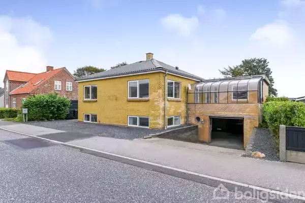 Gul murstensvilla med fladt tag og tilstødende carport ved vejen. Foran huset er der en grusbelagt indkørsel. Omgivet af hæk og andre huse i et villakvarter.