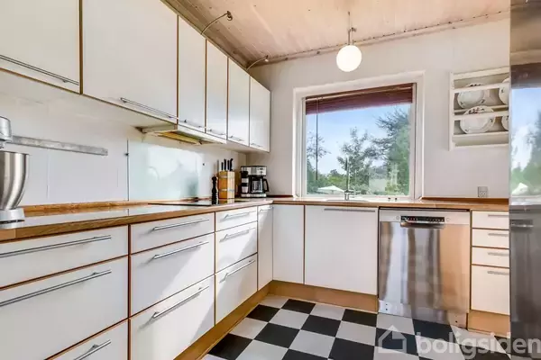 Et moderne køkken med hvidt træinteriør, en mixer på bordet og en opvaskemaskine under bordpladen. Rummet har et sort-hvidt skakternet gulv og udsigt til en have gennem vinduet.