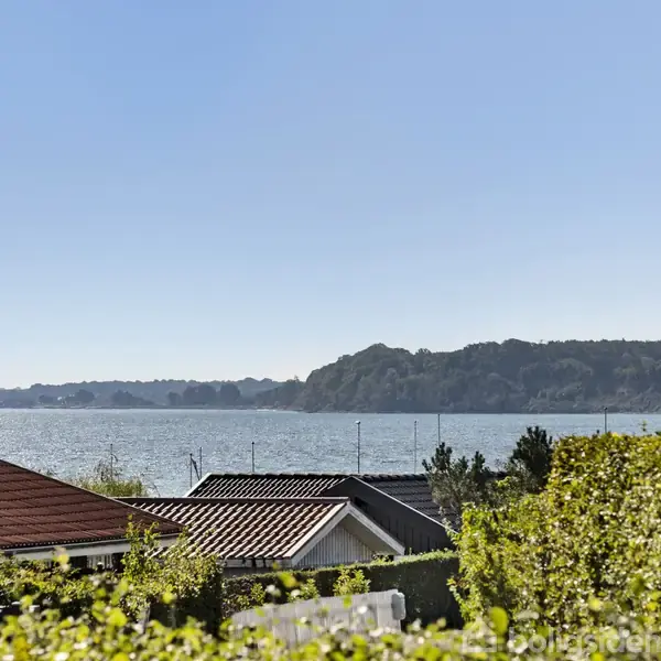 Røde hustage ses foran en blå sø med en skovbevokset kystlinje i baggrunden under en klar, blå himmel. Mellem husene står grønne buske.