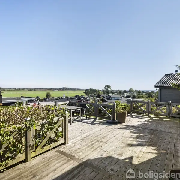 Stor træterrasse med pynteplanter på rækværket, hvor et bord og stole er placeret. I baggrunden ses en udsigt over marker og huse under en klar blå himmel.