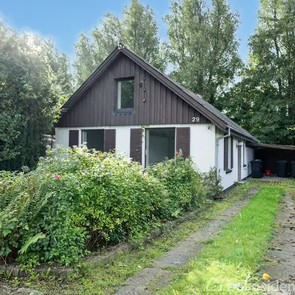 Hus med brune skodder og store vinduer står stille i en frodig have med grønne buske og træer, omkranset af en hæk. Klippeblok dekorerer plænen.