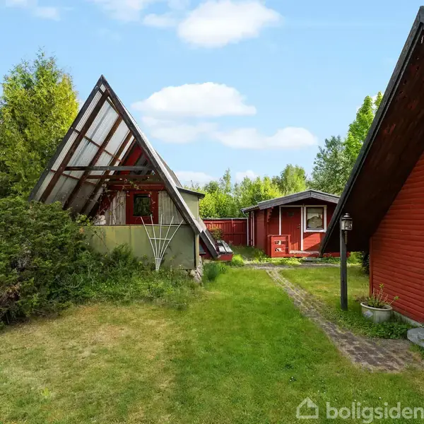 Rødt træhus med hvide dørkanter står i en grønt haveområde. Andet træhus med skråt tag ses i baggrunden. Grussti fører gennem græsset. Skov i horisonten.