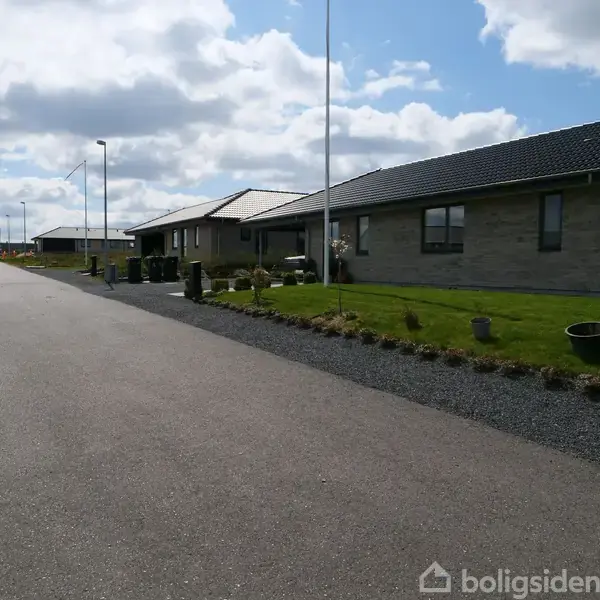 Hus med gråt murværk og mørkt tag står stille ved en velplejet have langs en stille vej under en skyet himmel.