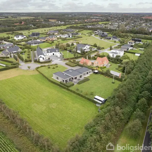 En ejendomskompleks ligger midt i et grønt, landligt område omgivet af andre huse. Veje og marker strækker sig gennem landskabet, mens træer flankerer en vej til højre.