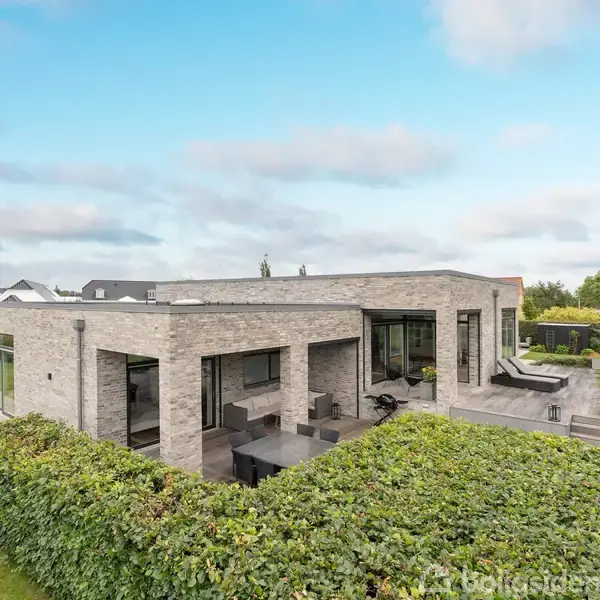 En moderne en-etagers murstensvilla med store vinduespartier står omgivet af en velplejet have med hække. Villaen har en terrasse, og omgivelserne er grønne med et klart, blåt skydække.