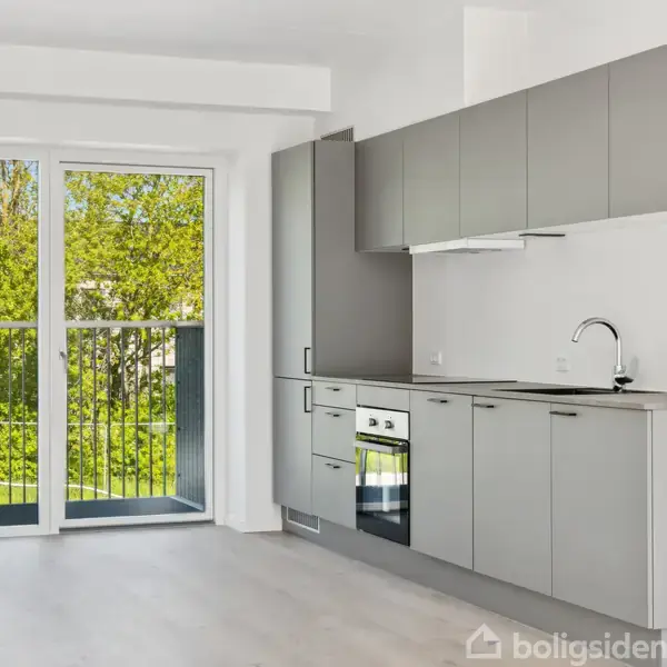 Et moderne køkken med grå skabe og indbygget ovn står stille i et lyst rum. Dobbetdør med glas fører ud til altan med udsigt til grønne træer.