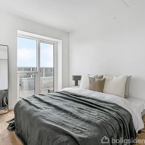 Soveværelse med en dobbeltseng dækket af en grå dyne og puder. Spejl står op ad væggen. Store vinduer giver udsigt til en balkon i en lys, minimalistisk indretning.