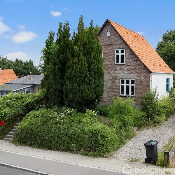 Et murstenshus med rødt tegltag står omgivet af frodige buske og træer ved en stille vej. Et fortov strækker sig langs huset, og en skraldespand er synlig.
