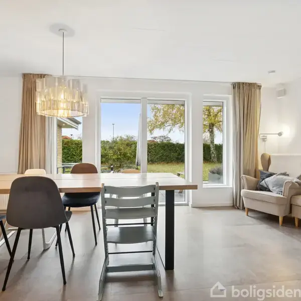 Et spisebord med stole står i et lyst rum. Store vinduer i baggrunden åbner ud til en have. En lænestol står ved siden af en bogreol til højre.