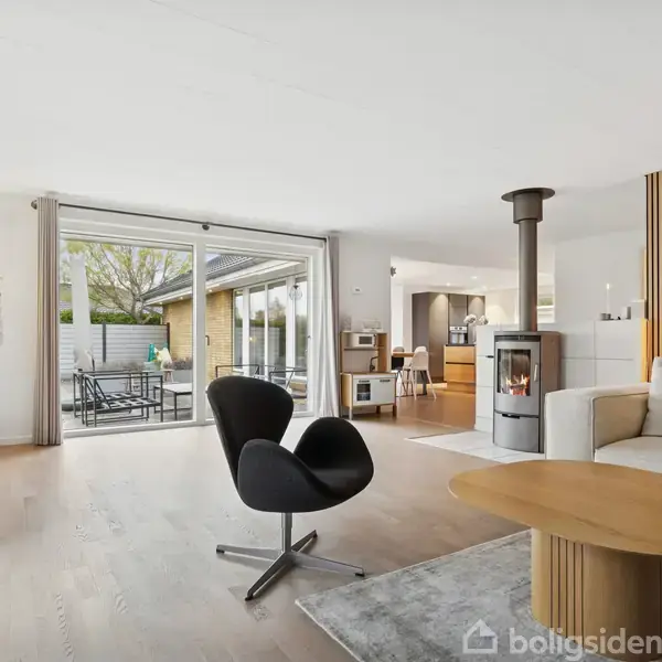 Stue med moderne lænestol foran sofabord. Pejs opvarmer rummet, der har lyse trægulve. Store glasdøre fører ud til terrasse med havemøbler, omgivet af hegn og have.