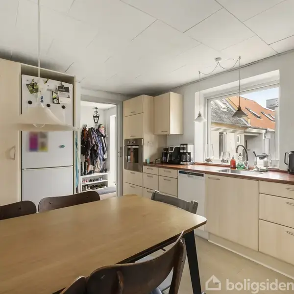 Et køkken med træbord omgivet af stole. Køleskabet står ved siden af skabene. Vindue med udsigt til hustage. Kaffe- og køkkenmaskiner på bordpladen.