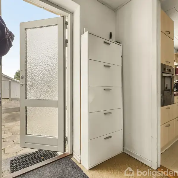 En åben dør står mod en terrasse, hvor jakker hænger til venstre. I højre del ses et hvidt skoskab og et køkken i baggrunden med lyse træskabe.