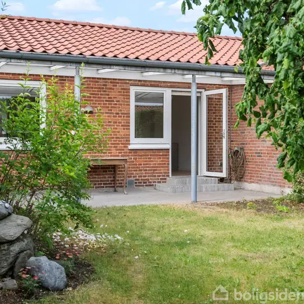 Murstenshus med rødt tegltag, står med åben dør til terrasse. Omkranset af græsplæne, buske og træer i en frodig have.