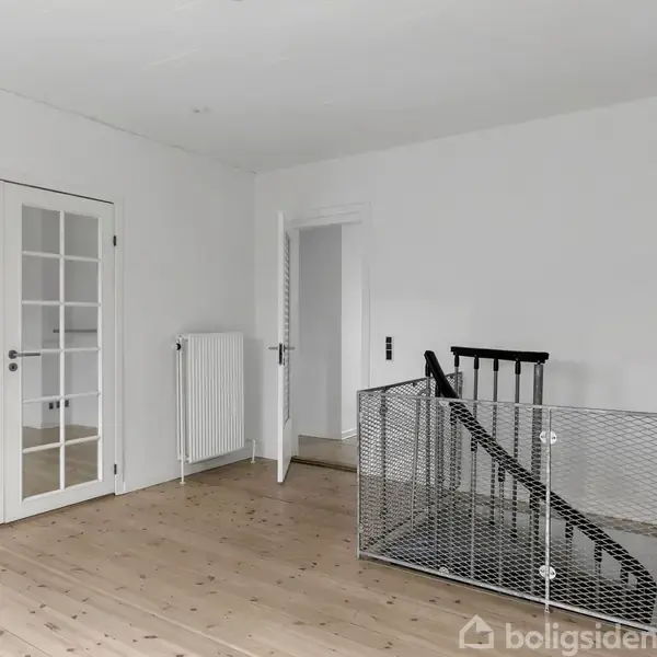 Stue med trægulve, franske døre og en trappe med sort gelænder. Væggene er hvide, og der er en radiator ved en åben dør til et andet rum.