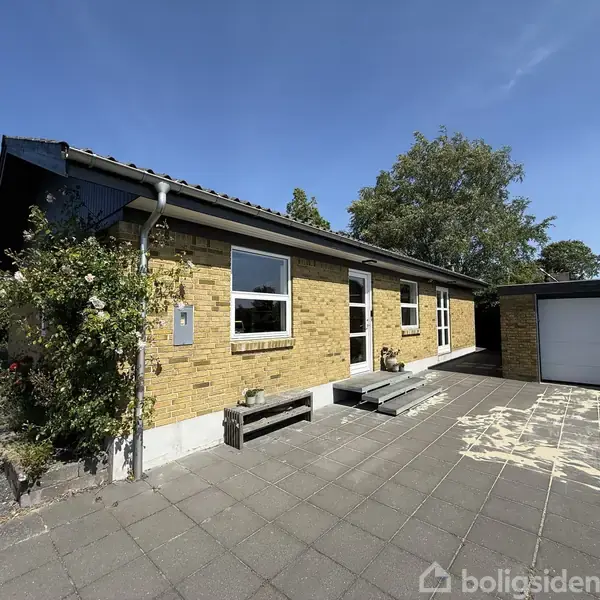 En gul murstensvilla står stationær på en flisebelagt gårdsplads med tilstødende garage; omgivet af en frodig have med træer under en klar blå himmel.
