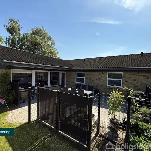 Hus med brun murstensfacade omgivet af en have og terrasseområde, afskærmet med et hegn. Overdækket terrasse rummer møbler. Foran huset står en solsikke. Tekst: "Visualisering".