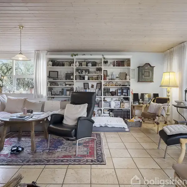 Stue med sofa og lænestol omkring et sofabord på et tæppe. Bøger fylder en reol i baggrunden. Rummet har flisegulv, store vinduer og flere lamper.