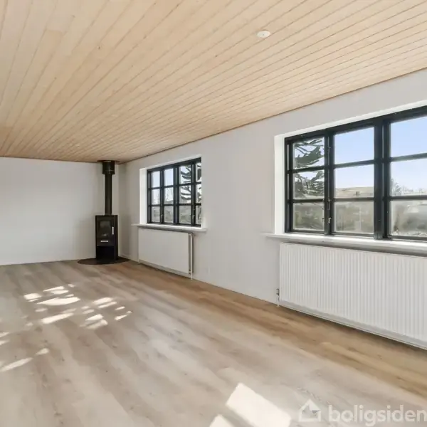 Stue med træloft og trægulv, store vinduer med sort ramme, brændeovn i hjørnet, hvide vægge og radiatorer. Rummeligt og lyst.