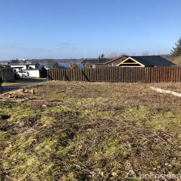 En åben grund med spredt græs og visne grene ligger i forgrunden, mens et træhegn omkranser grunden. Flere mindre huse og en vej ses i baggrunden.