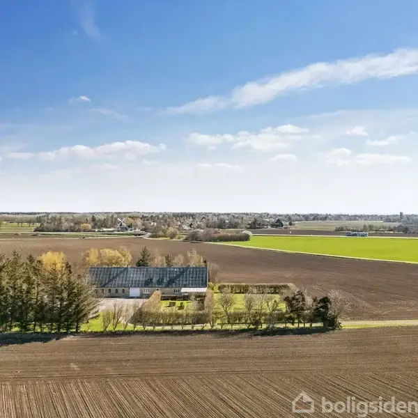 Et hus står stille blandt marker, omgivet af træer. I baggrunden ses spredte bebyggelser og vidtstrakte landbrugsarealer under en klar himmel.
