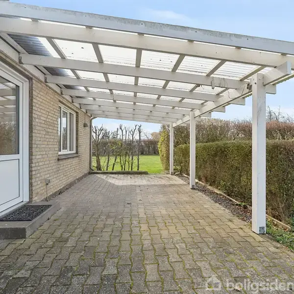Terrasse med træpergola dækker flisebelagt område ved murstenshus. Græsplæne og buskads omgiver terrassen i en rolig have.