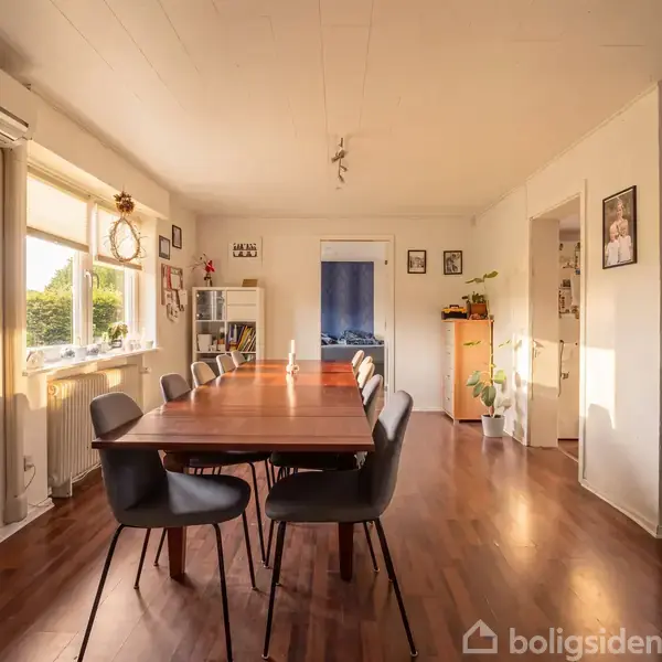 Spisebord med sorte stole står centralt. Sollys strømmer ind gennem vinduet til venstre. Væggene er dekoreret med billeder. Til højre er en sofa og planter i hjørnet.
