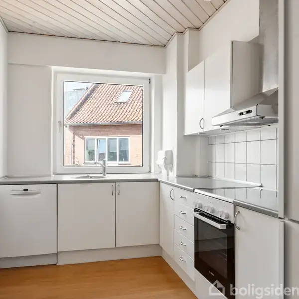 Et hvidt, moderne køkken med L-formet bordplade. Der er en ovn, emhætte og opvaskemaskine. Et vindue over vasken viser taget af en nabobygning.