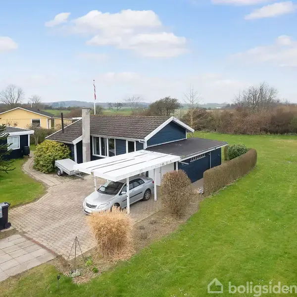 Et blå hus med hvidt tag og carport står på en grøn grund med omkringliggende buske. En bil er parkeret i indkørslen. Flagstang i baggrunden.