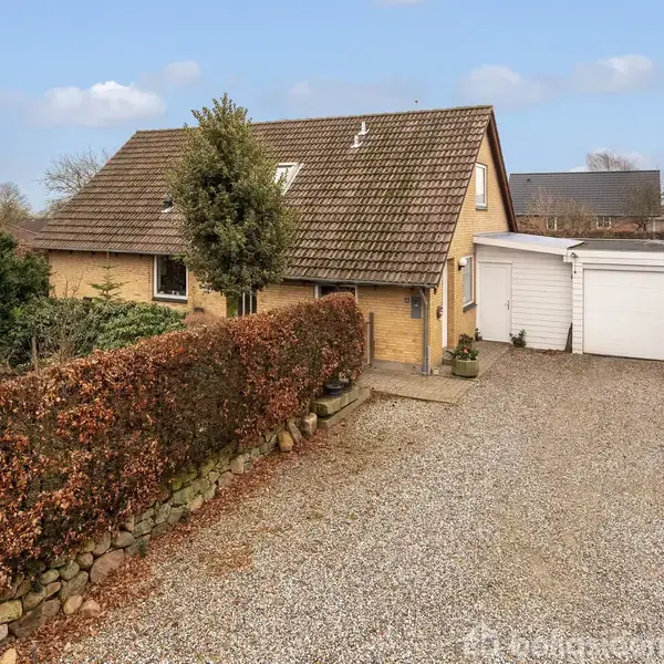 Et gult murstenshus med skråtag står i en have omkranset af hække. Huset har en grusindkørsel og en hvid garage ved siden af. Et skilt med teksten "home" er placeret ved indkørslen.