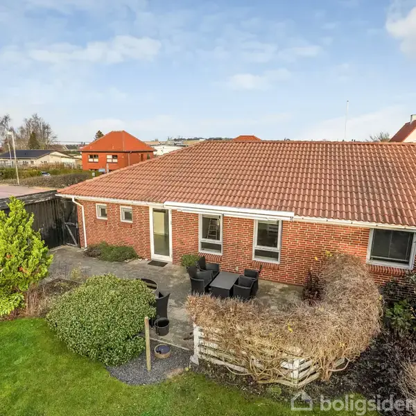 Murstenshus med rødt tegltag står i en grøn have med fliser, buske og havemøbler. Andre huse ses i baggrunden under en blå himmel.