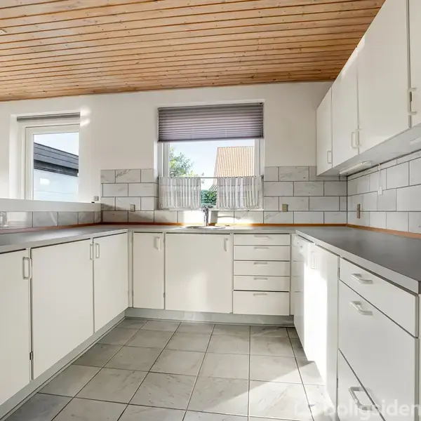 Et moderne køkken med hvide skabe og skuffer er placeret i et lyst rum. Fliser på væggene, et vindue med dagslys og en træloft kompletterer indretningen.