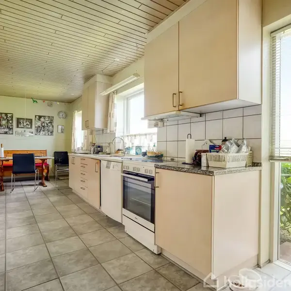 Køkken med komfur, skabsplads, og opvask i gang. Spisebord og stole står i baggrunden ved vindue, der giver udsigt til have. Tæppebelagt gulv og træbeklædt loft.