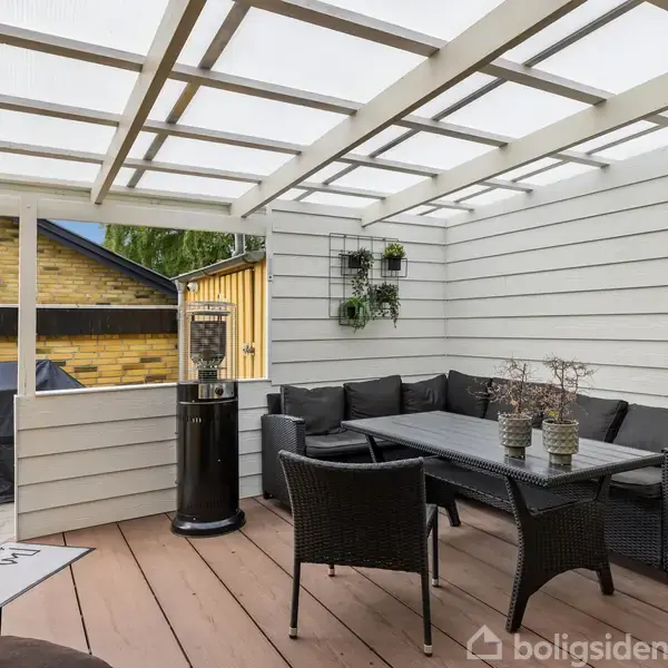 Terrasseområde med grå sofa og sorte stole omkring et bord. Overdækningen er gennemsigtig, og væggen er hvidmalet. Udenfor ses en grill dækket med et sort overtræk.