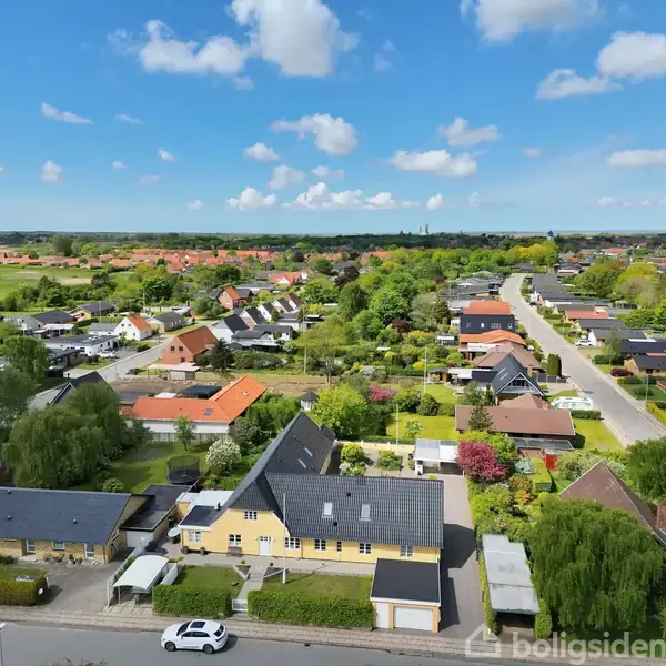 Rækkehuse i en parcelhuskvarter med haver, træer og grønne områder. Veje løber gennem kvarteret under en klar blå himmel med få skyer, der skaber en fredelig stemning.
