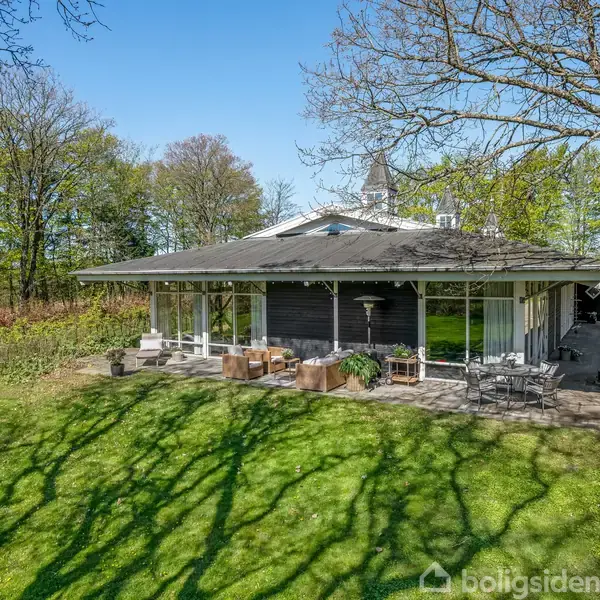 Et moderne hus med fladt tag og store glaspartier, står fredfyldt i en grøn have omkranset af træer. Terrassen er møbleret med siddepladser, og skygger fra træer falder på græsset.