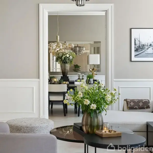 Lys og elegant stue med gråt sofamøblement. Friske blomster på et sofabord. I baggrunden ses en spisestue med et spisebord dekoreret med blomster. Væggene er lyse med en sort-hvid fotografi.